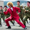 A Japanese comedian with wavy blonde hair, wearing a red suit, actively participating in a Japanese Self-Defense Forces (JSDF) training session. The scene shows the comedian in military gear, engaging in a physical drill alongside JSDF soldiers in an outdoor training ground.